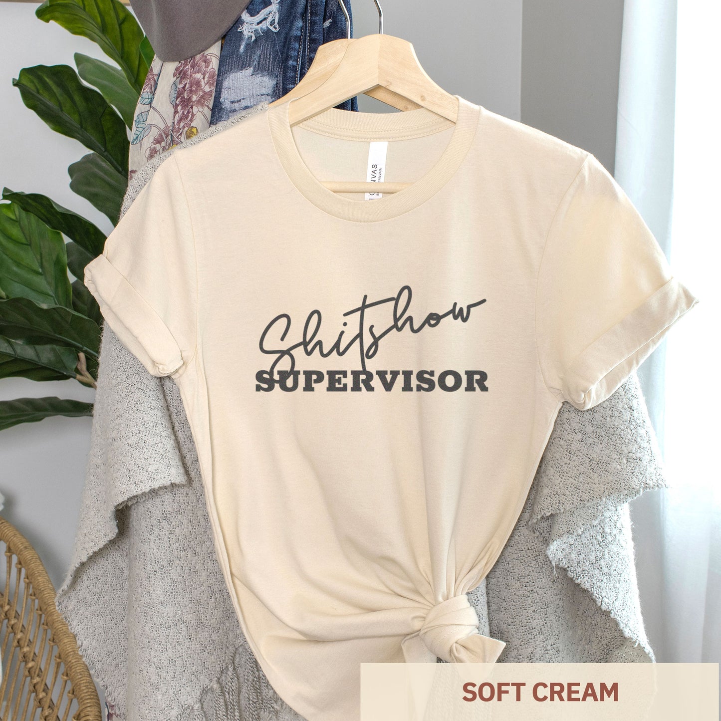 A hanging soft cream Bella Canvas t-shirt featuring the text shitshow supervisor.