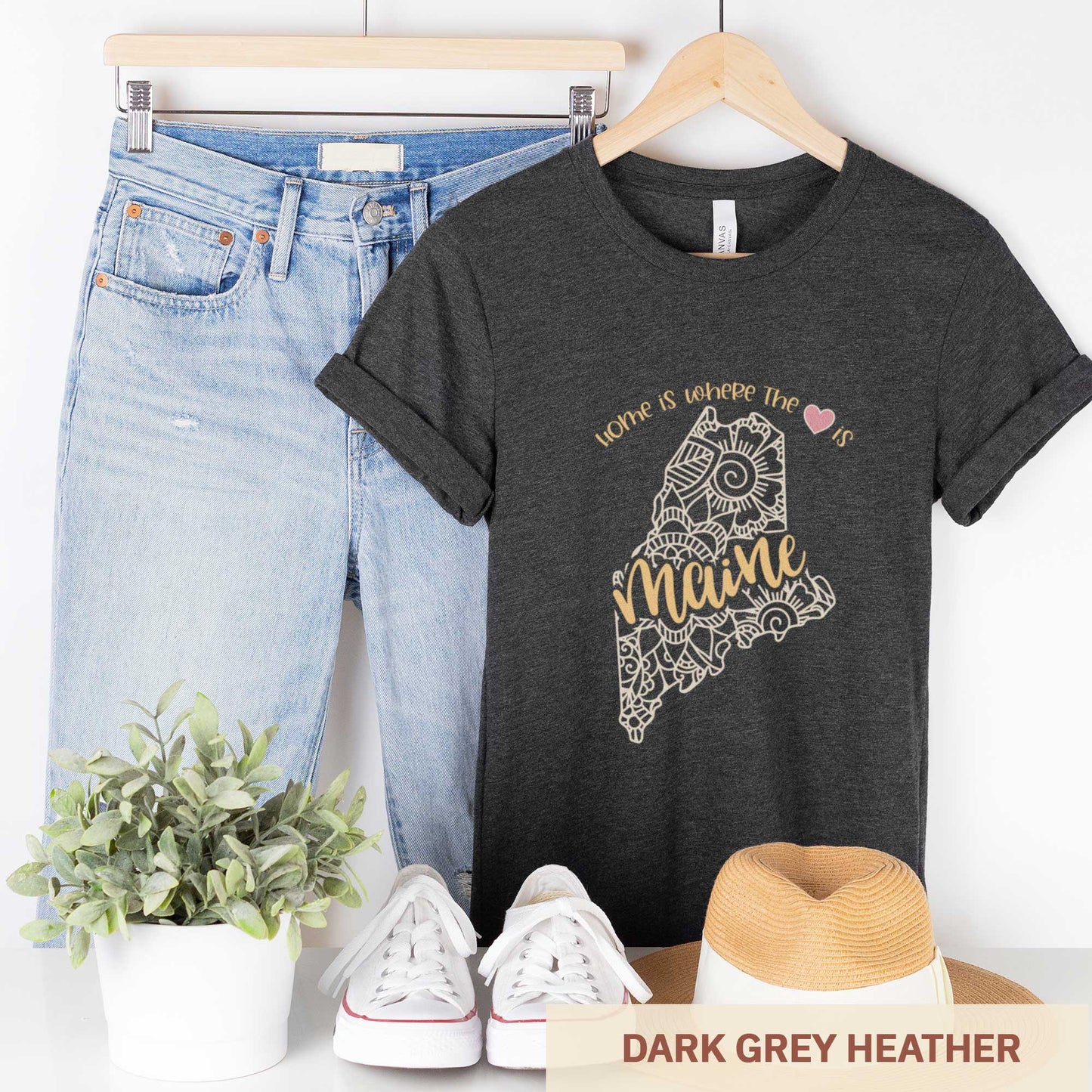 A hanging dark grey heather Bella Canvas t-shirt featuring a mandala in the shape of Maine with the words home is where the heart is.