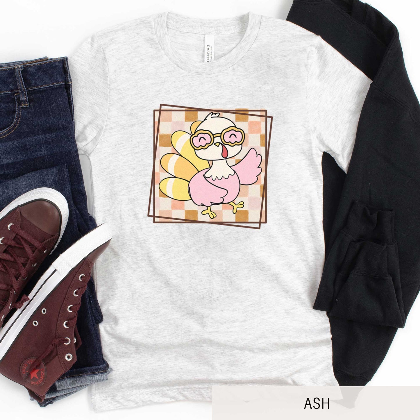 An ash Bella Canvas t-shirt featuring a cute cartoon turkey against a fall checkered background.