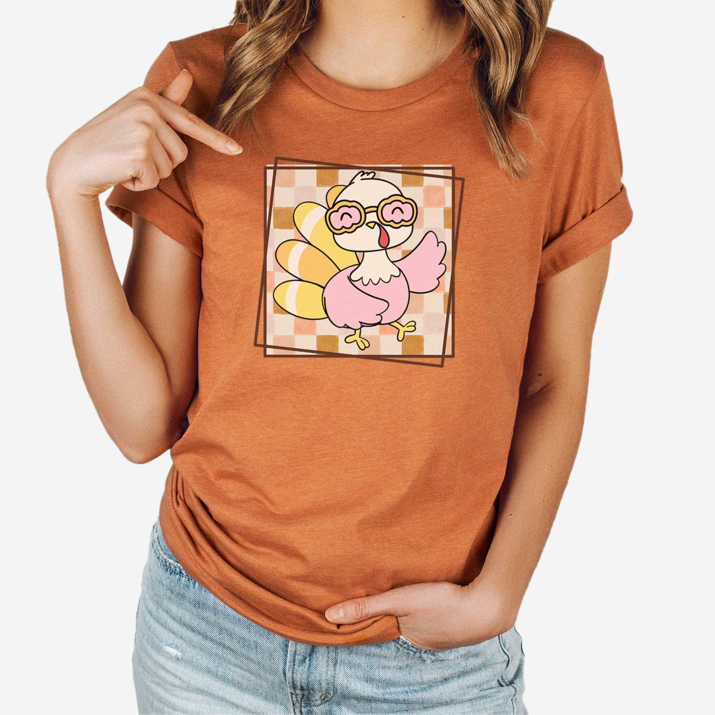 A woman wearing a heather autumn Bella Canvas t-shirt featuring a cute cartoon turkey against a fall checkered background.