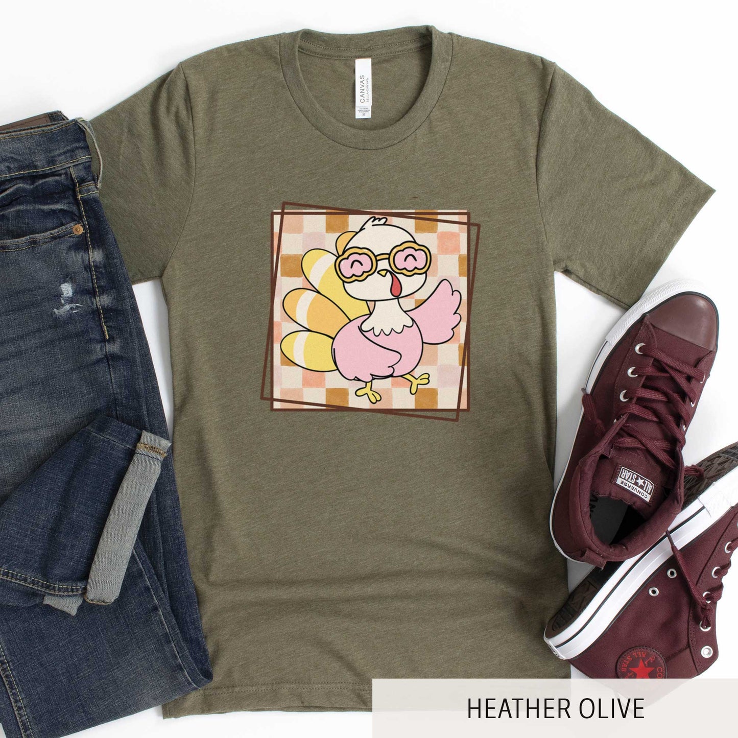 A heather olive Bella Canvas t-shirt featuring a cute cartoon turkey against a fall checkered background.
