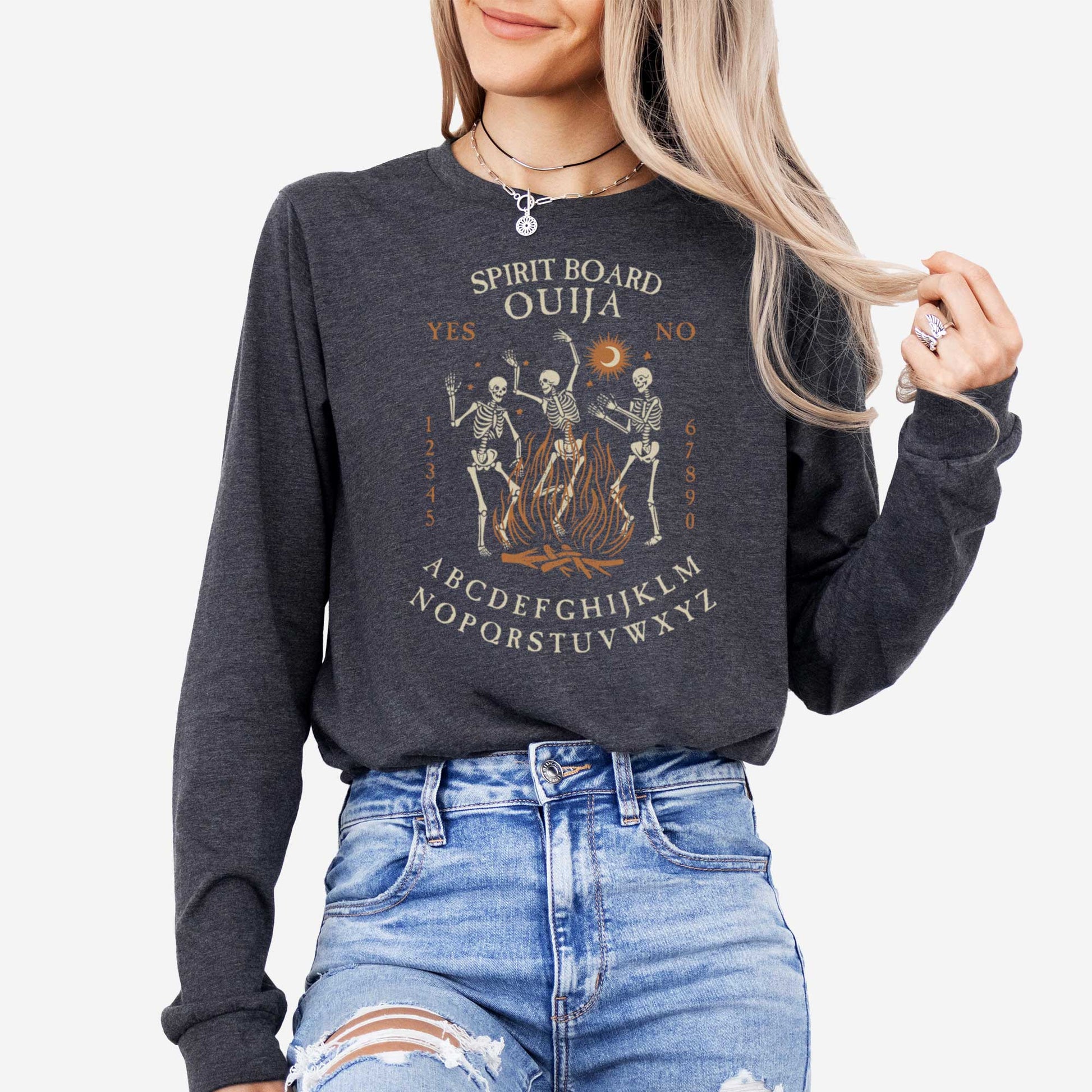 A woman wearing a dark grey heather Bella Canvas long sleeve t-shirt with dancing skeletons in front of a fire and an ouija board layout.