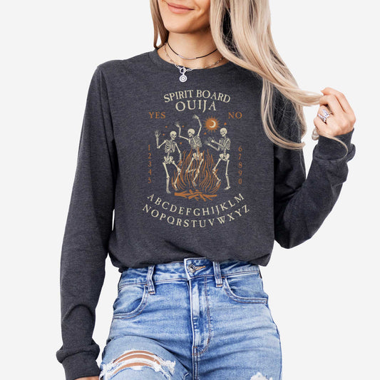 A woman wearing a dark grey heather Bella Canvas long sleeve t-shirt with dancing skeletons in front of a fire and an ouija board layout.