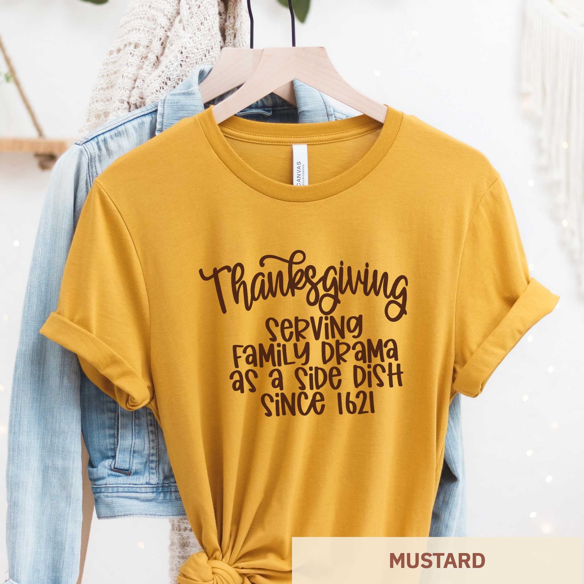 A hanging mustard yellow Bella Canvas t-shirt featuring the words thanksgiving serving family as a side dish since 1621.