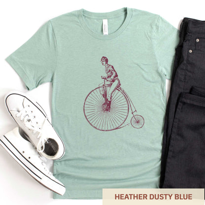 An heather dusty blue Bella Canvas t-shirt featuring a Victorian man riding a penny farthing bicycle.