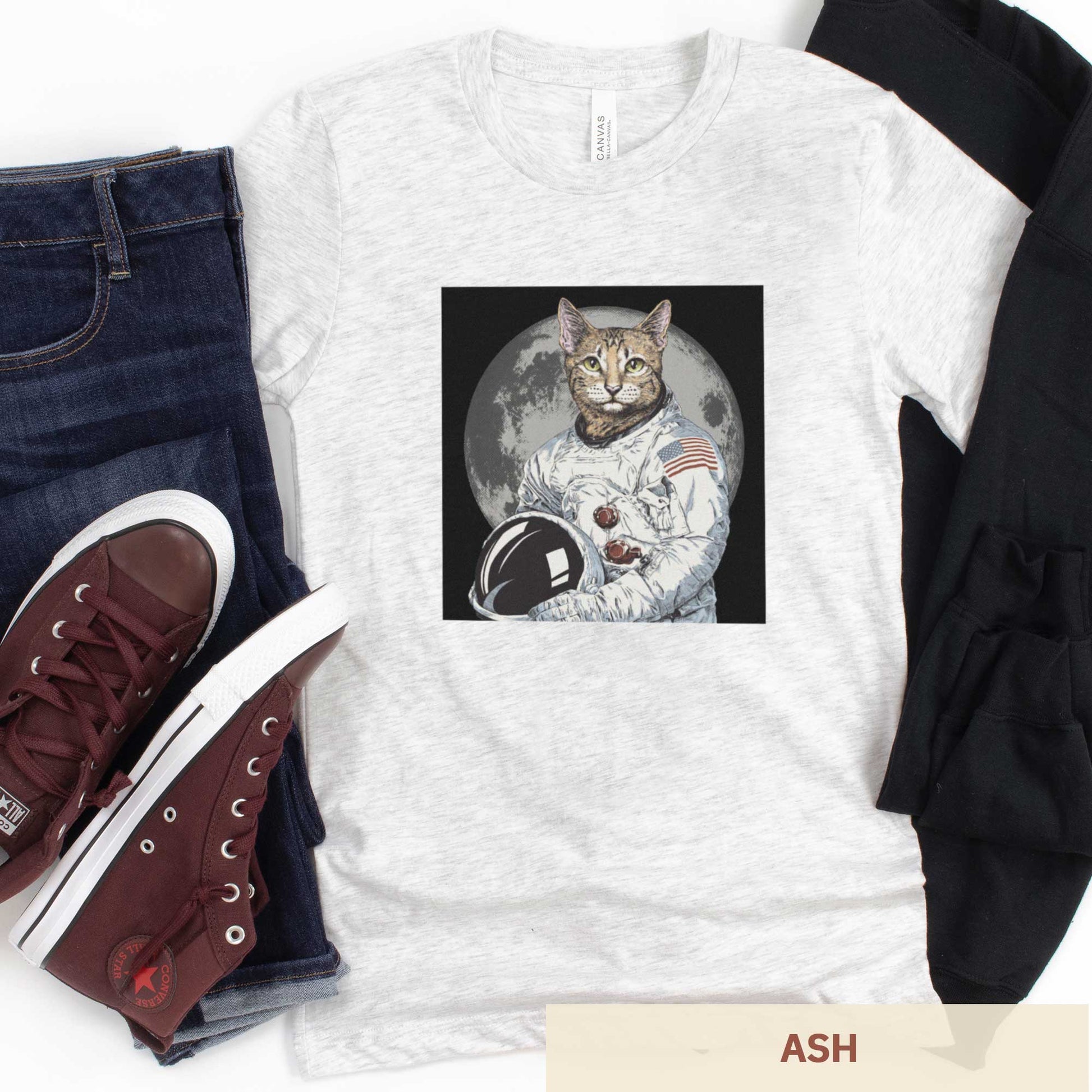 An ash Bella Canvas t-shirt featuring an illustrated portrait of a cat as a NASA astronaut with the moon in the background.
