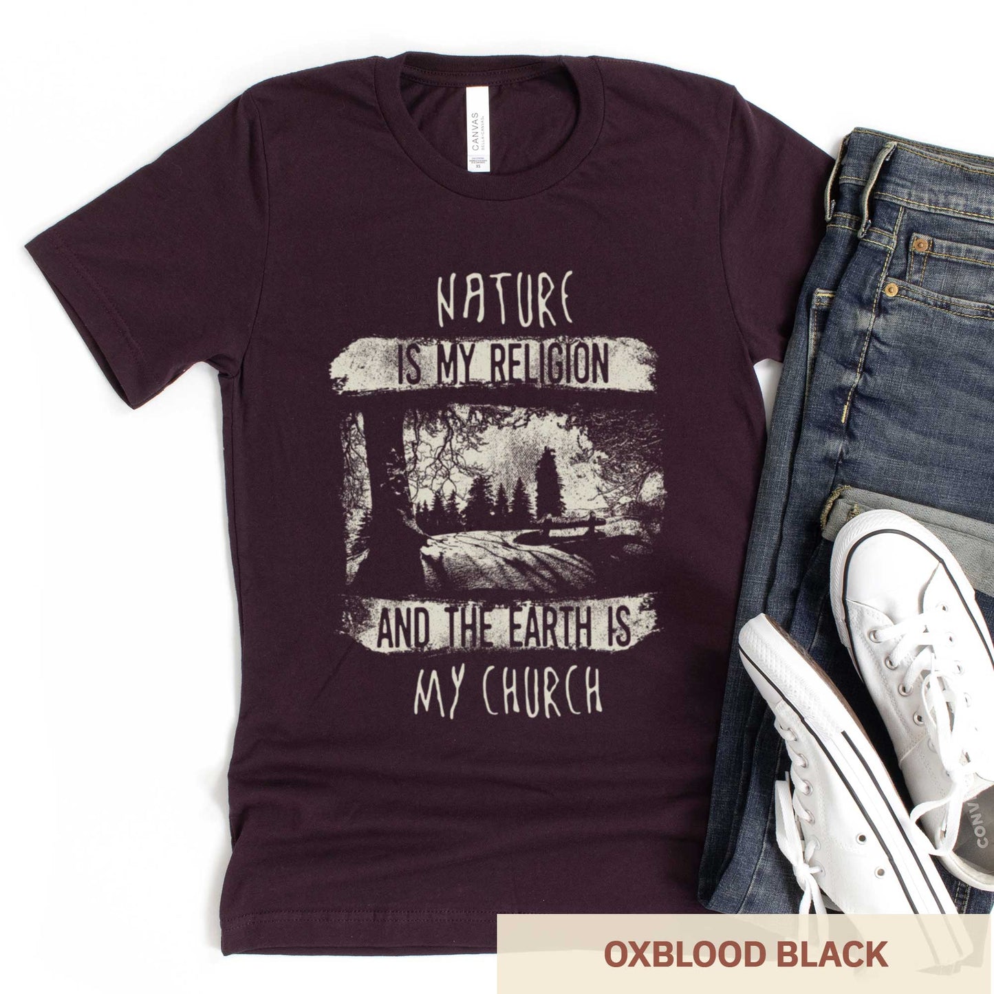 An oxblood black Bella Canvas t-shirt featuring fields and a forest with the words nature is my religion and the earth is my church.