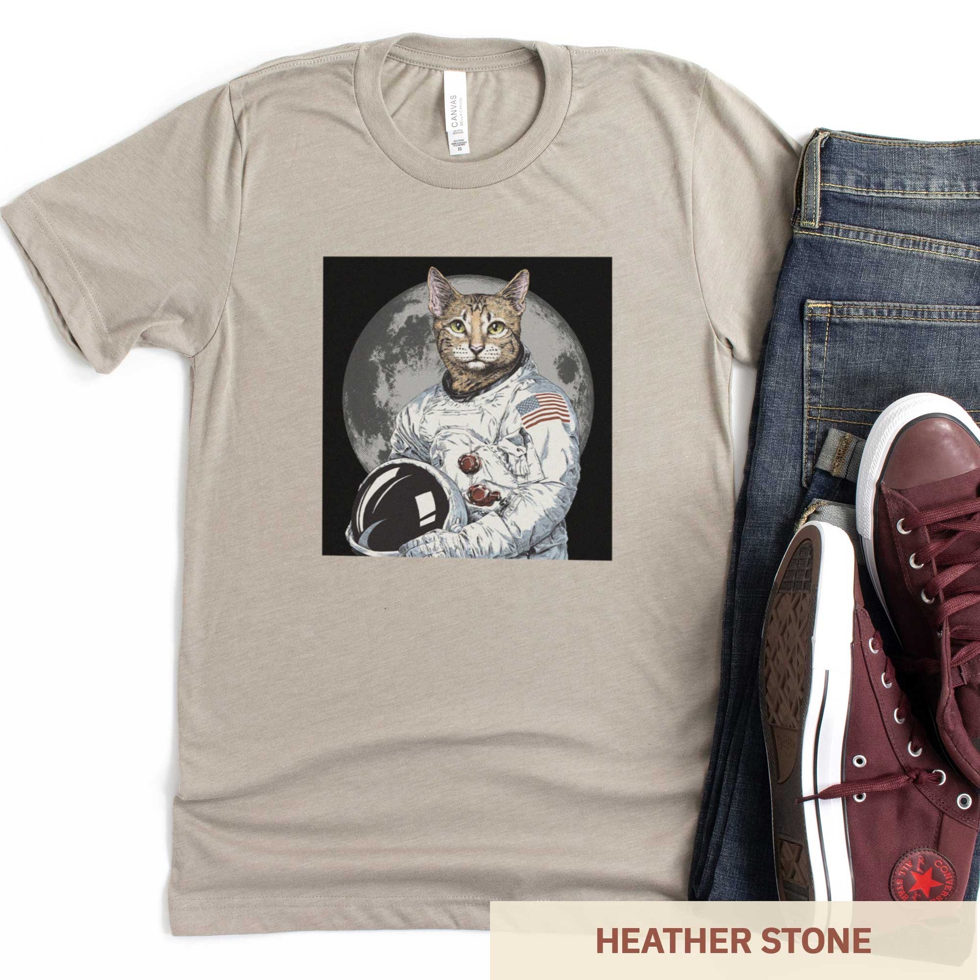 A heather stone Bella Canvas t-shirt featuring an illustrated portrait of a cat as a NASA astronaut with the moon in the background.