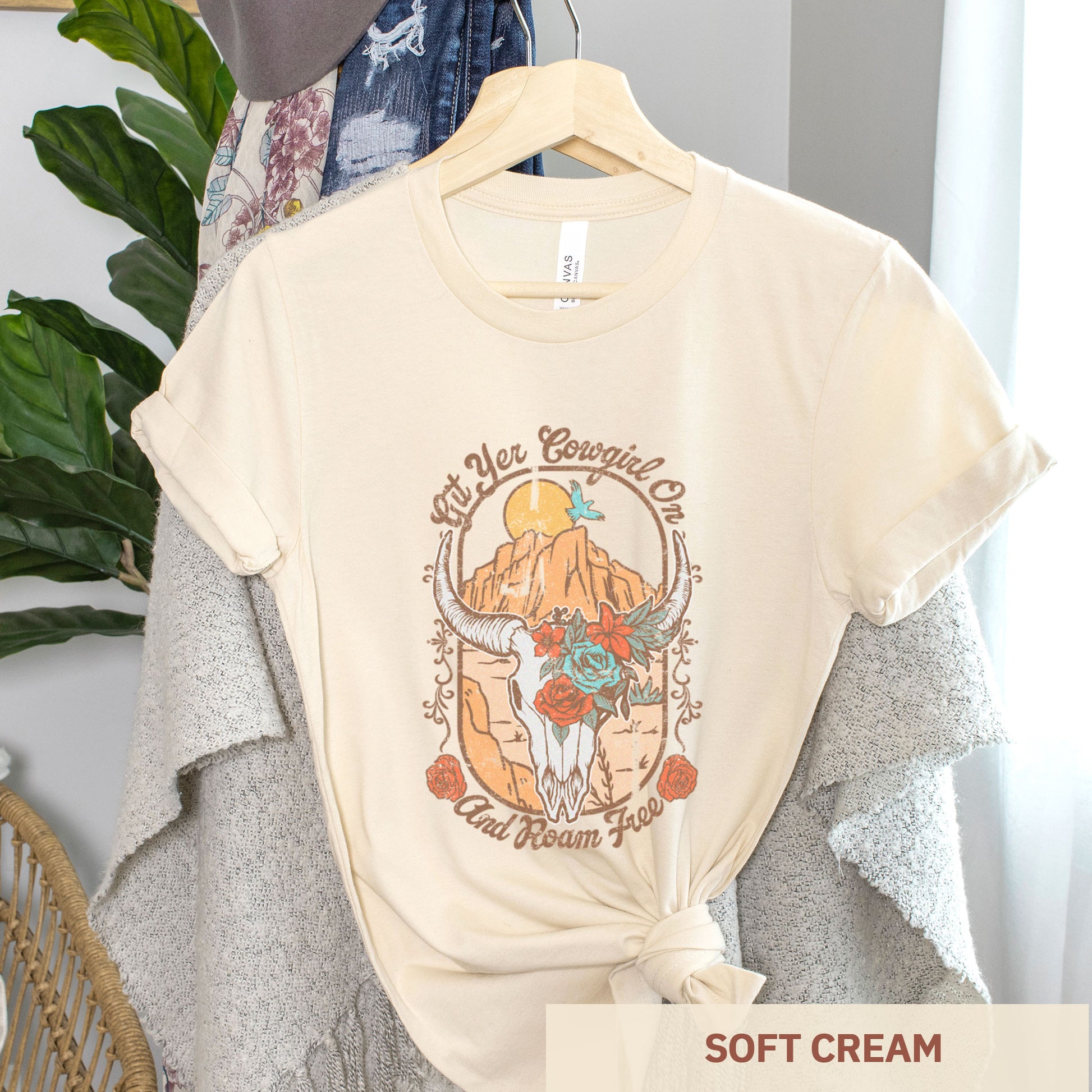 A hanging soft cream peach Bella Canvas t-shirt featuring a cow skull that says git yer cowgirl on.