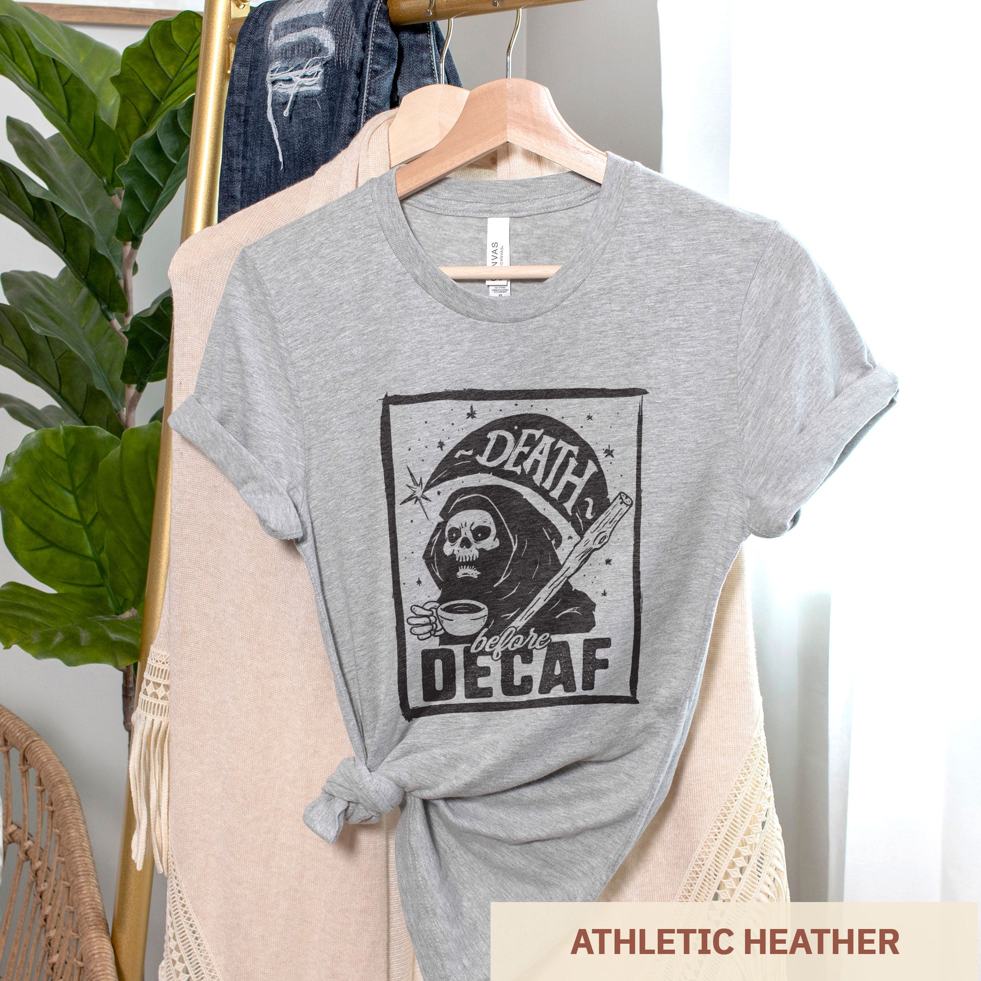 A hanging athletic heather Bella Canvas t-shirt featuring the grim reaper drinking a cup of coffee with the words Death before decaf
