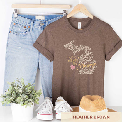 A hanging heather brown Bella Canvas t-shirt featuring a mandala in the shape of Michigan with the words home is where the heart is.