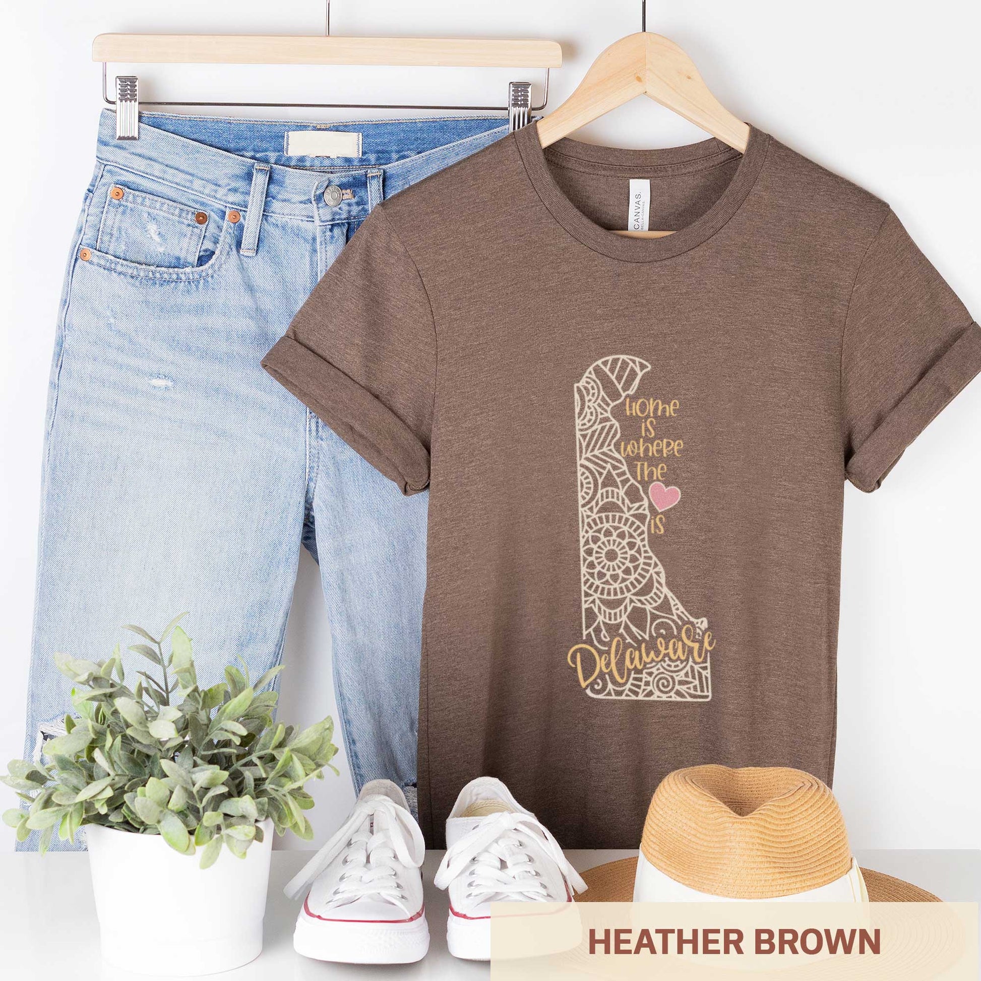 A hanging heather brown Bella Canvas t-shirt featuring a mandala in the shape of Delaware with the words home is where the heart is.