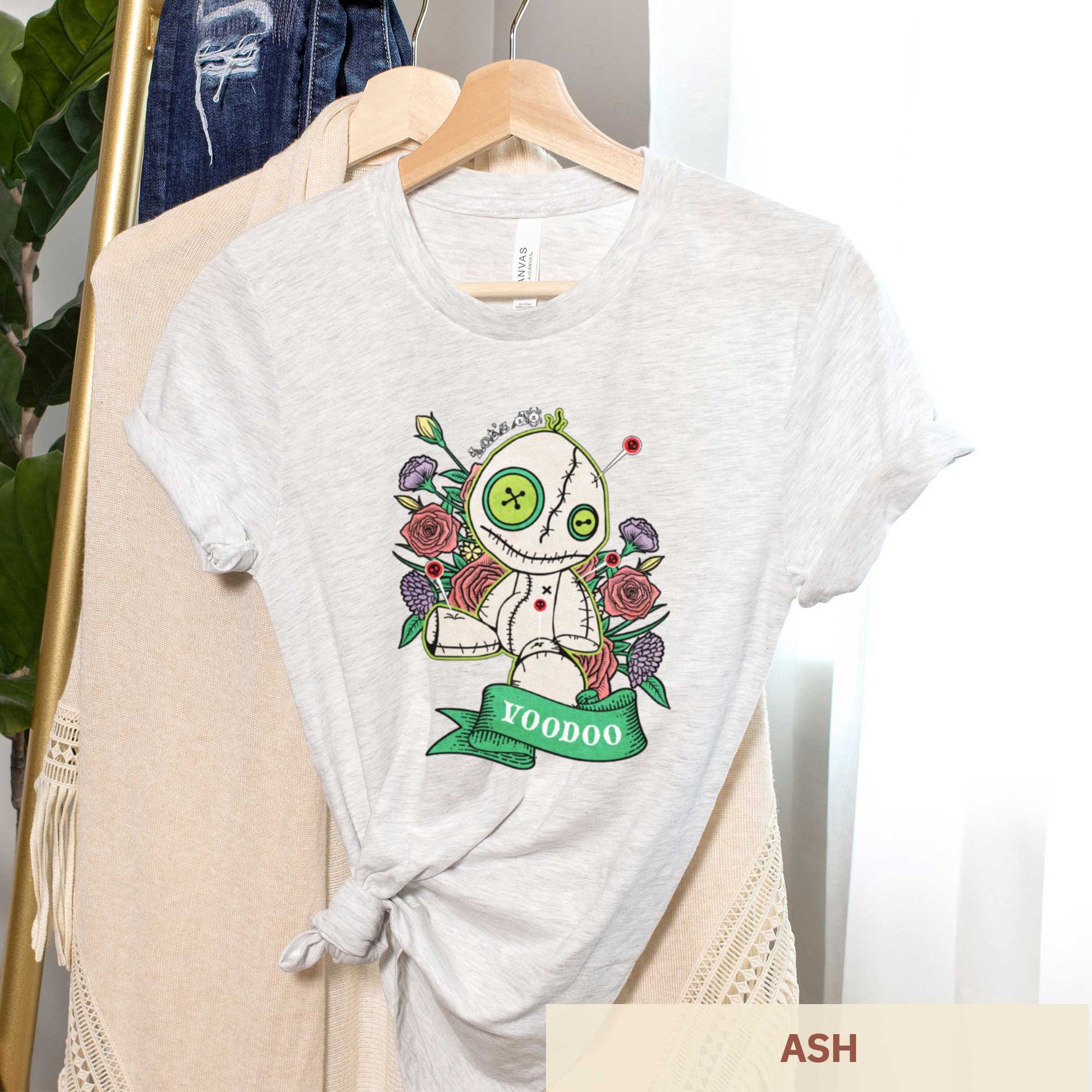 An hanging ash Bella Canvas t-shirt featuring a cartoon voodoo doll and flowers.
