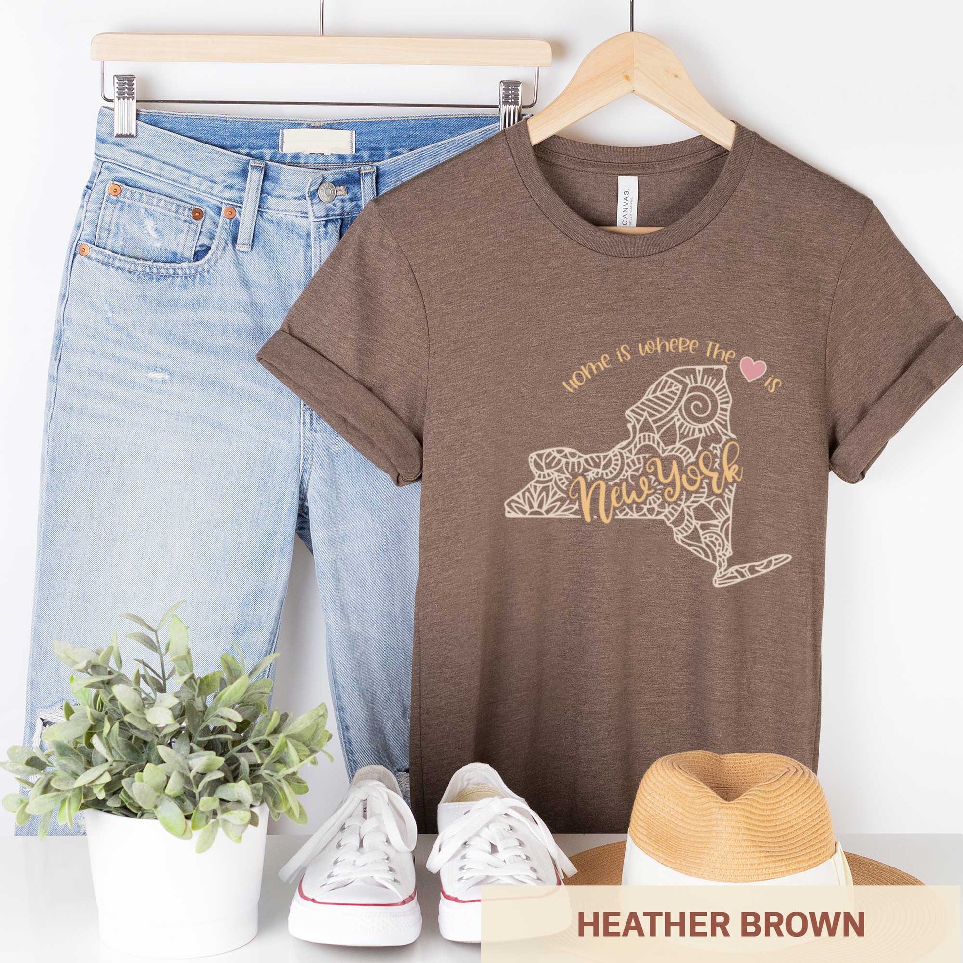 A hanging heather brown Bella Canvas t-shirt featuring a mandala in the shape of New York with the words home is where the heart is.