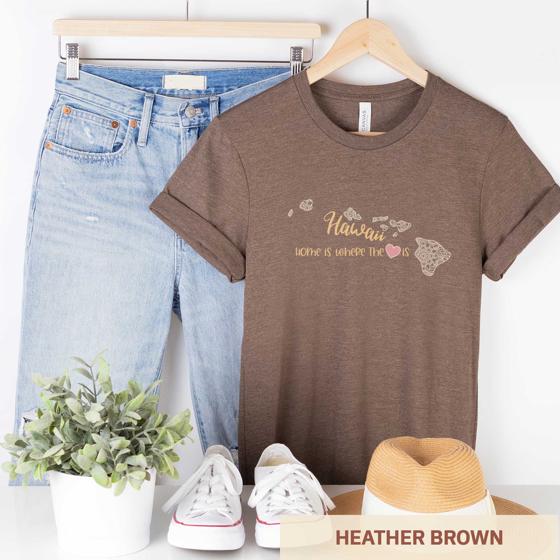 A hanging heather brown Bella Canvas t-shirt featuring a mandala in the shape of Hawaii with the words home is where the heart is.