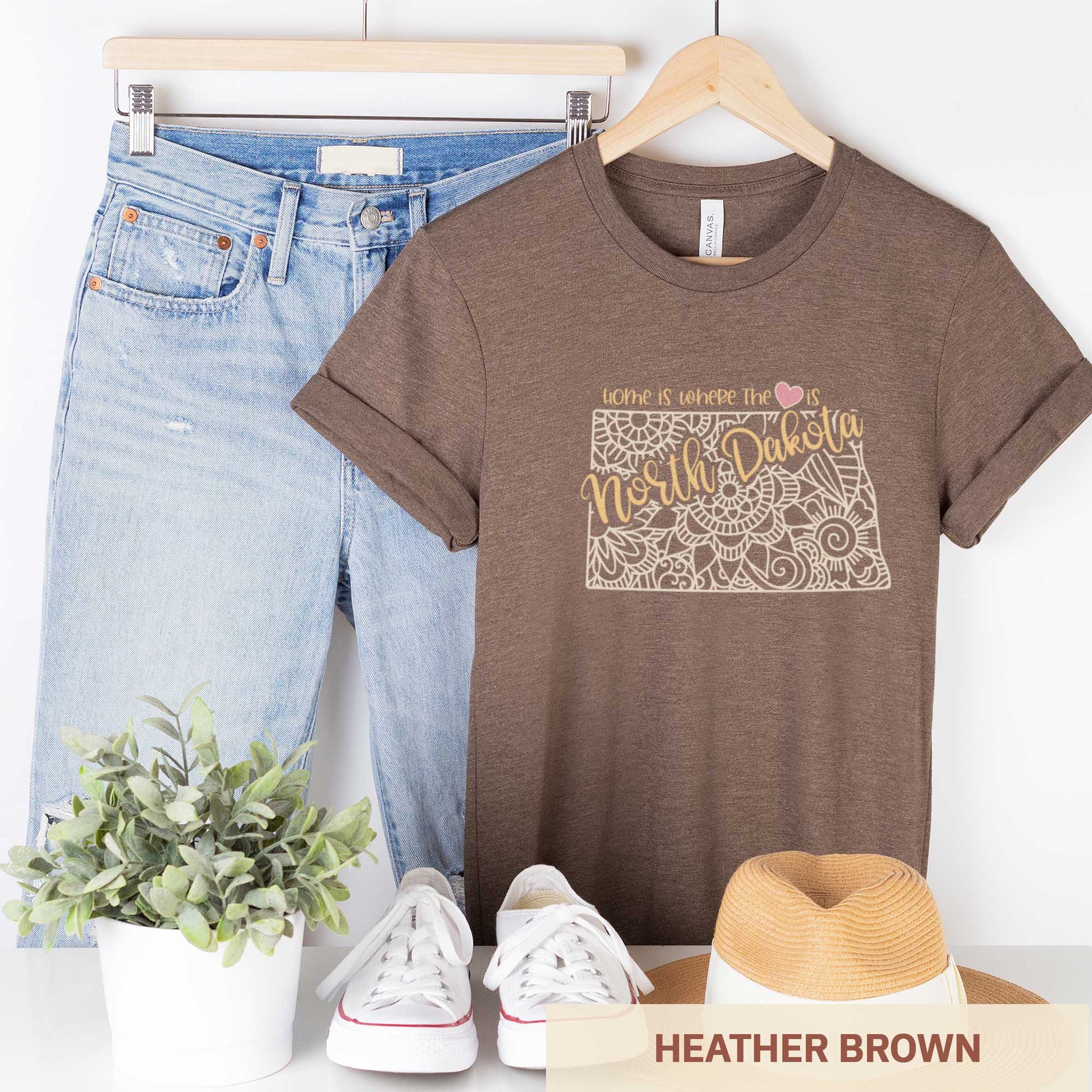 A hanging heather brown Bella Canvas t-shirt featuring a mandala in the shape of North Dakota with the words home is where the heart is.