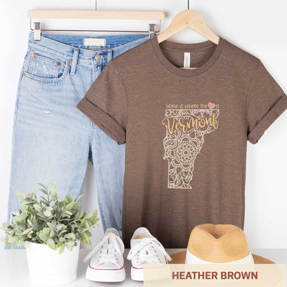 A hanging heather brown Bella Canvas t-shirt featuring a mandala in the shape of Vermont with the words home is where the heart is.