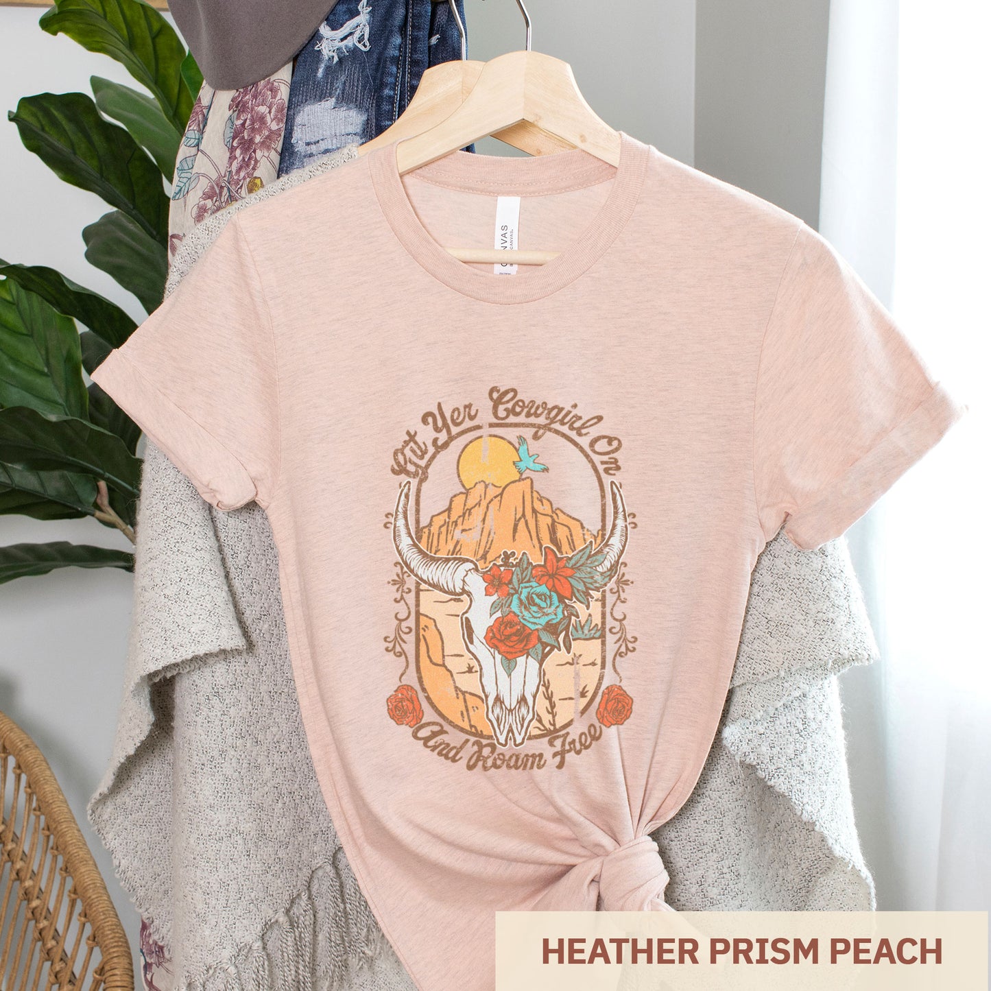 A hanging heather prism peach Bella Canvas t-shirt featuring a cow skull that says git yer cowgirl on.