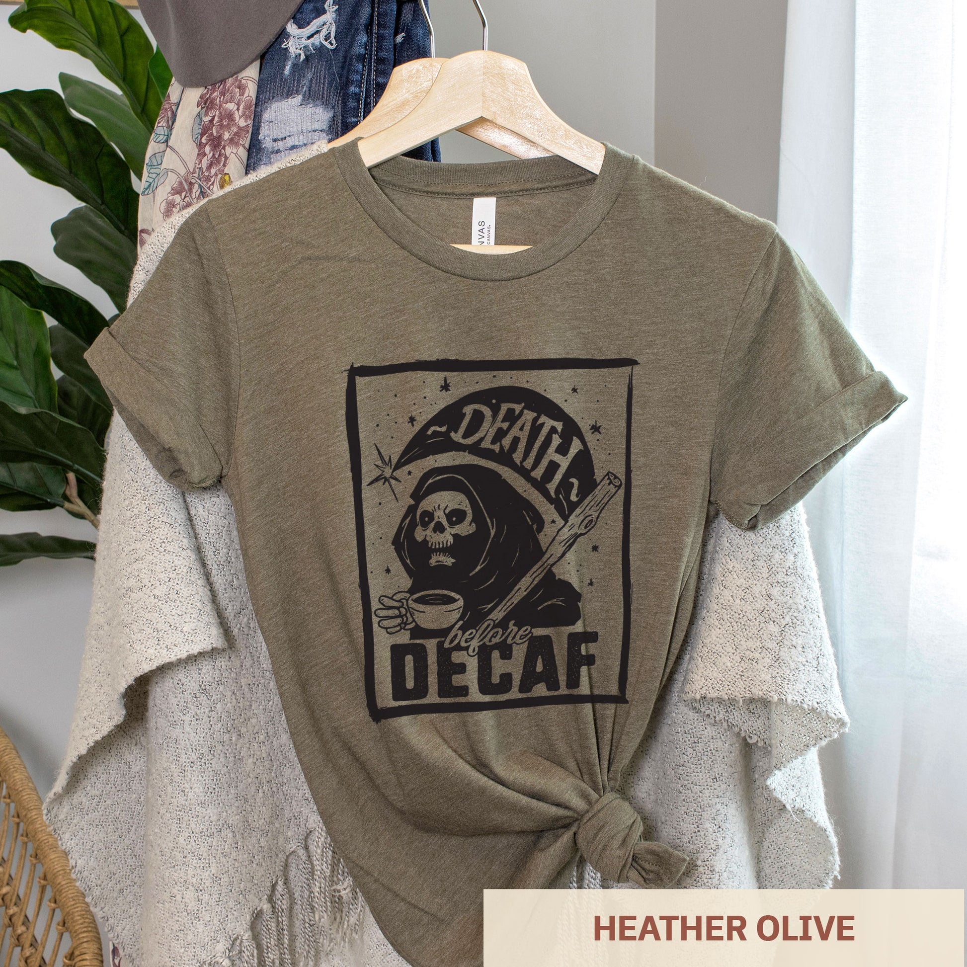 A hanging heather olive Bella Canvas t-shirt featuring the grim reaper drinking a cup of coffee with the words Death before decaf
