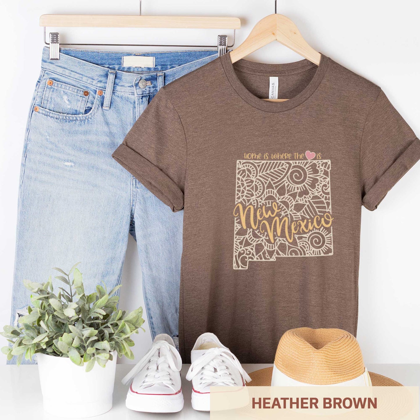A hanging heather brown Bella Canvas t-shirt featuring a mandala in the shape of New Mexico with the words home is where the heart is.