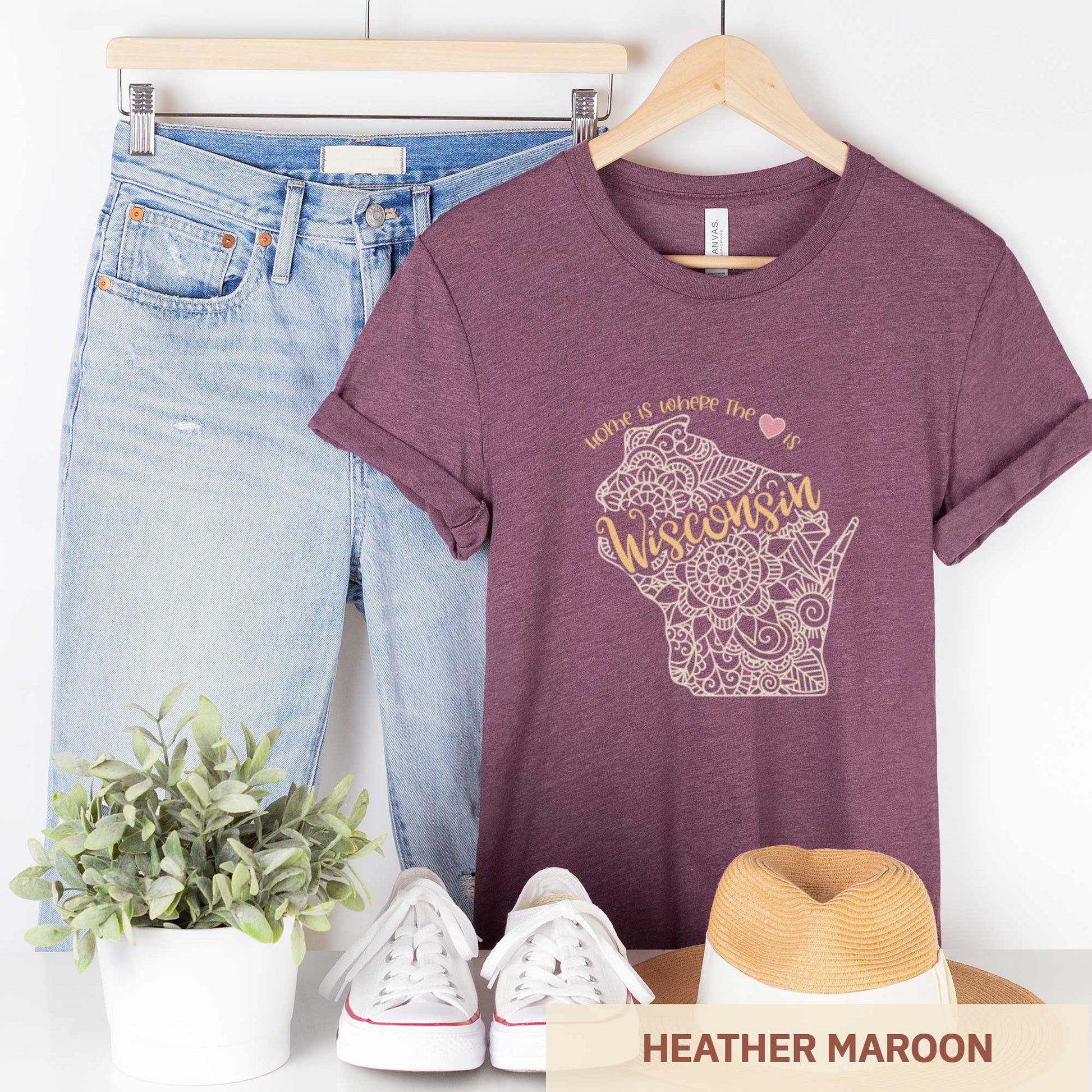 A hanging heather maroon Bella Canvas t-shirt featuring a mandala in the shape of Wisconsin with the words home is where the heart is.