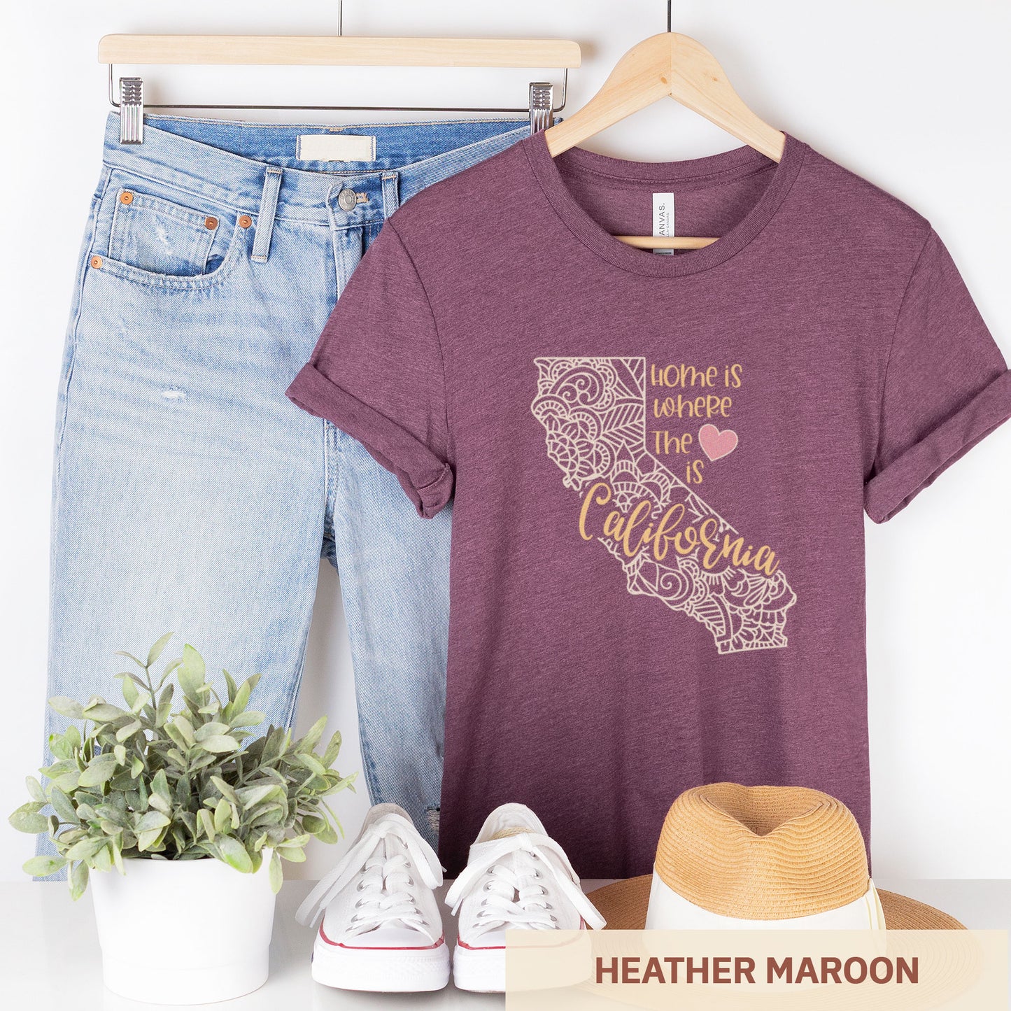 A hanging heather maroon Bella Canvas t-shirt featuring a mandala in the shape of California with the words home is where the heart is.