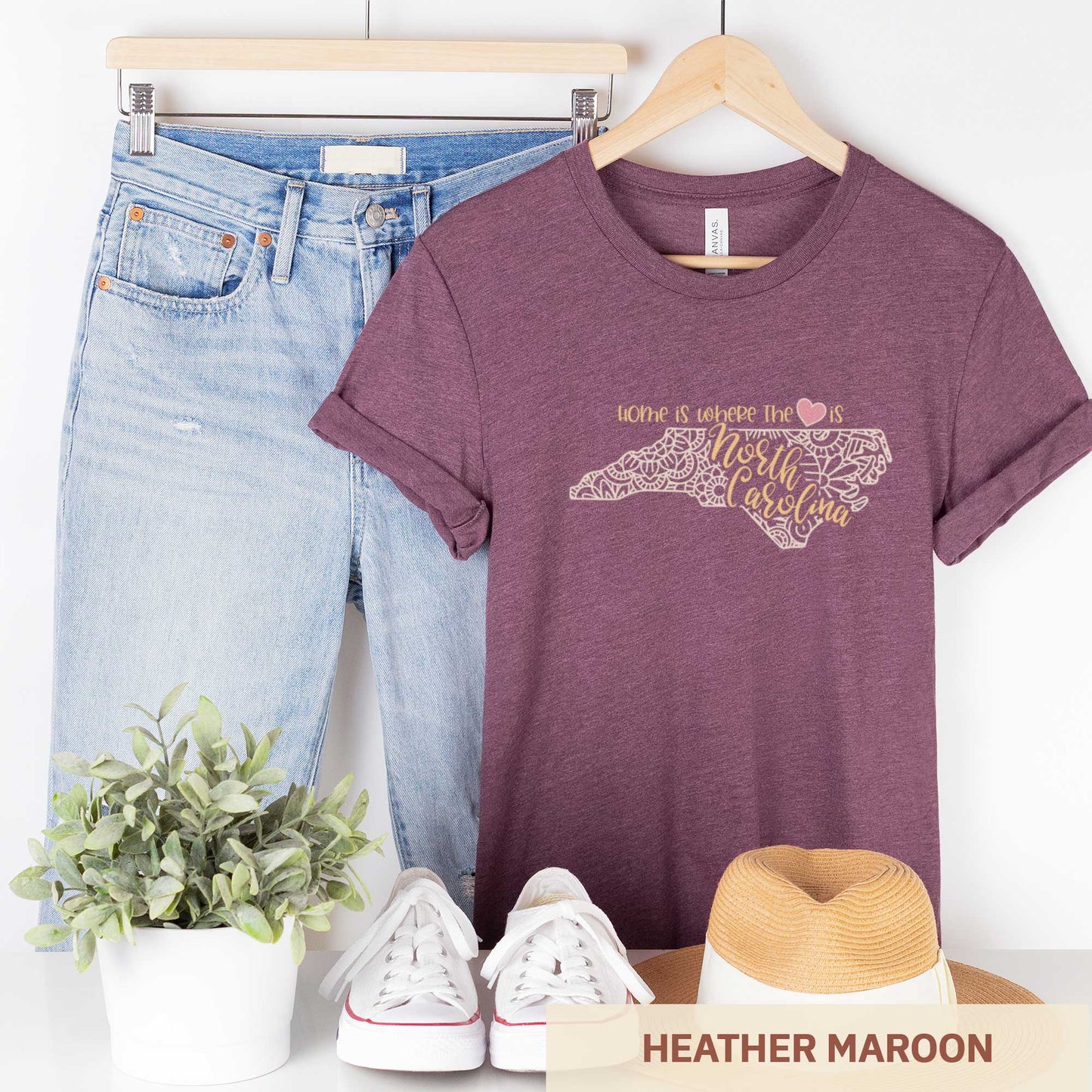 A hanging heather maroon Bella Canvas t-shirt featuring a mandala in the shape of North Carolina with the words home is where the heart is.