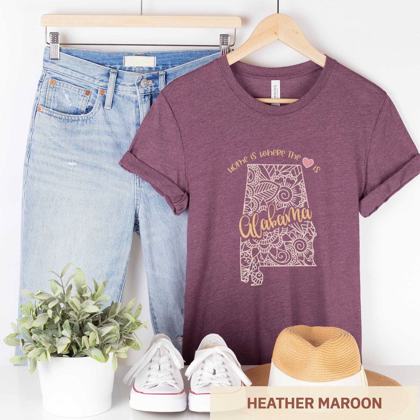 A hanging heather maroon Bella Canvas t-shirt featuring a mandala in the shape of Alabama with the words home is where the heart is.