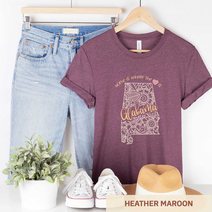 A hanging heather maroon Bella Canvas t-shirt featuring a mandala in the shape of Alabama with the words home is where the heart is.