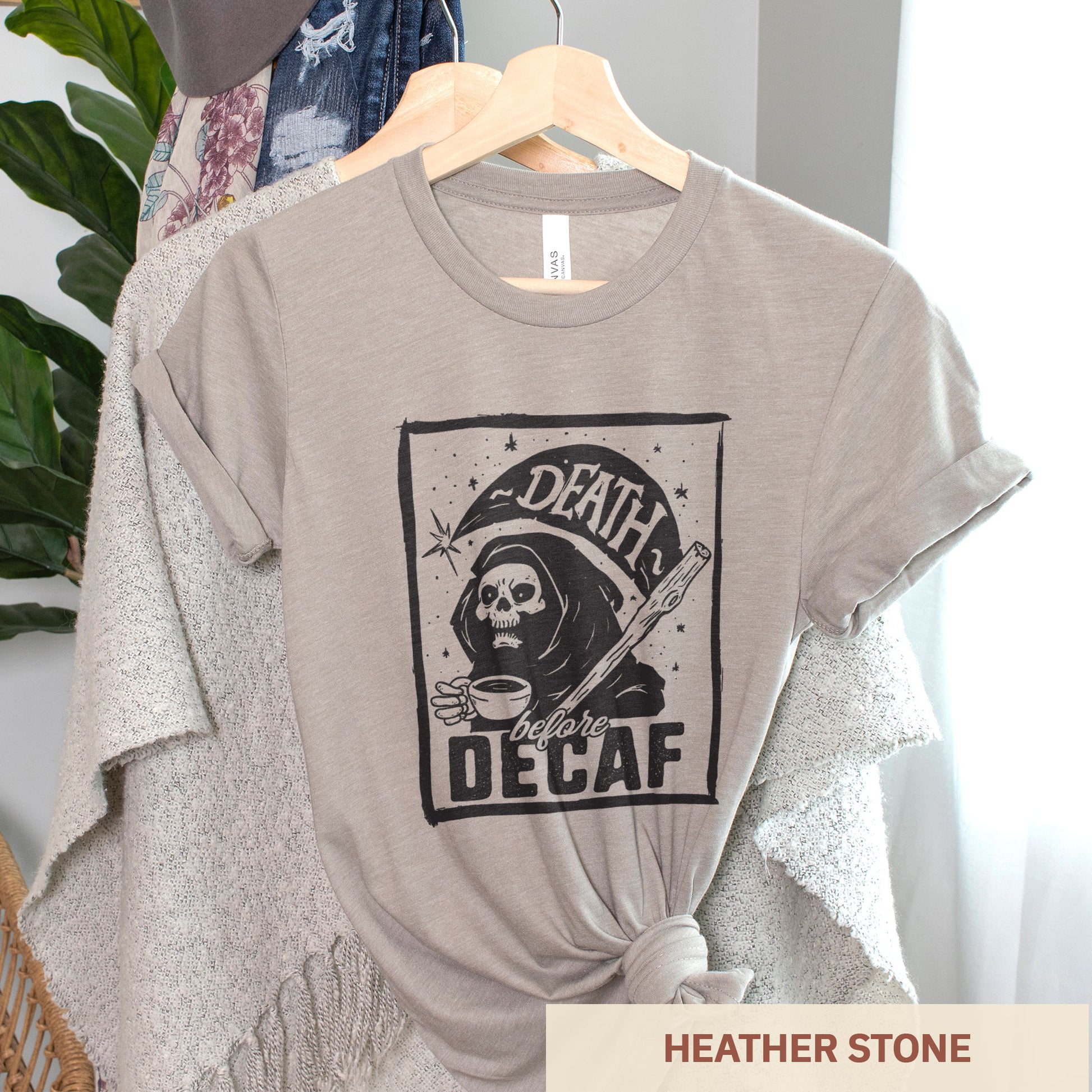 A hanging heather stone Bella Canvas t-shirt featuring the grim reaper drinking a cup of coffee with the words Death before decaf