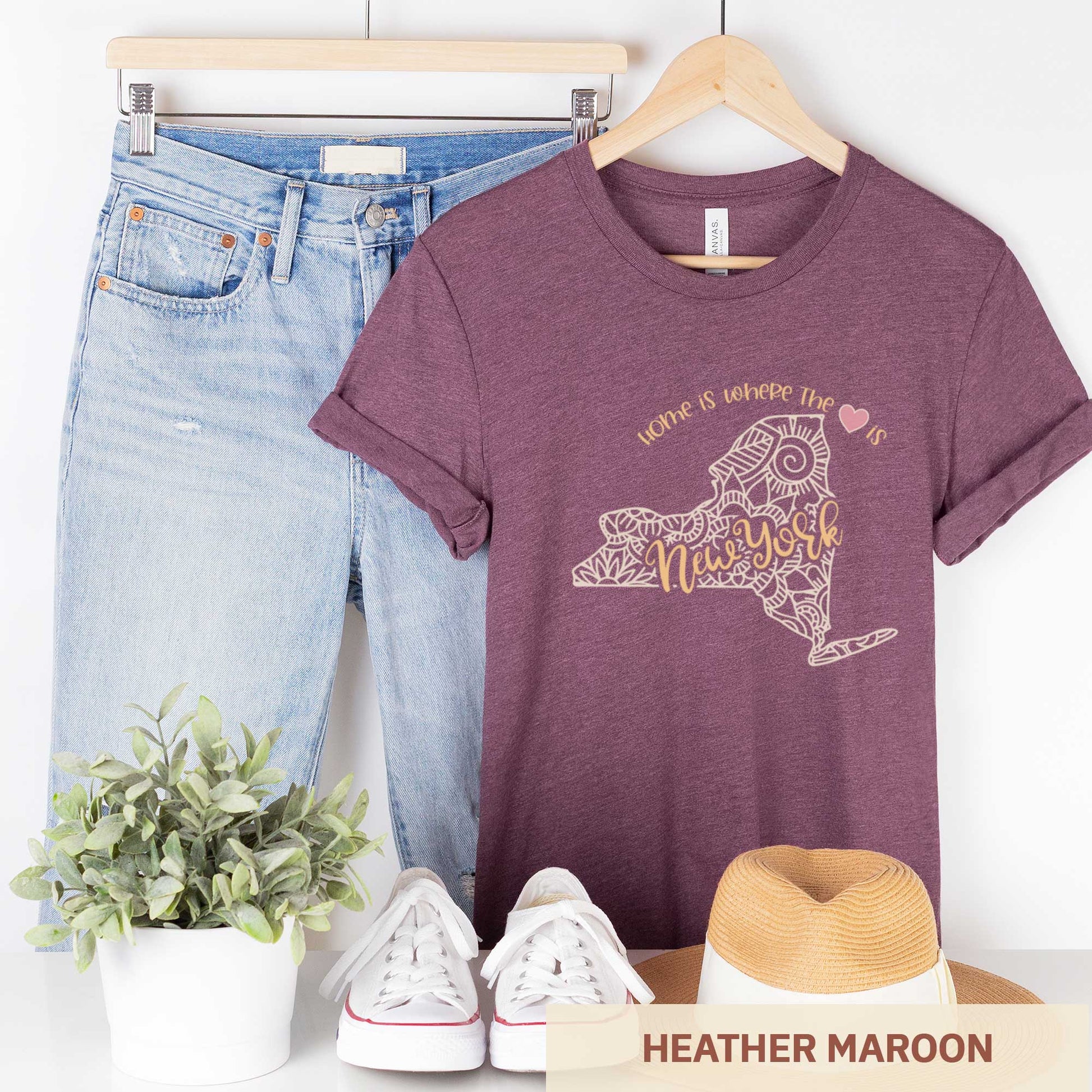 A hanging heather maroon Bella Canvas t-shirt featuring a mandala in the shape of New York with the words home is where the heart is.