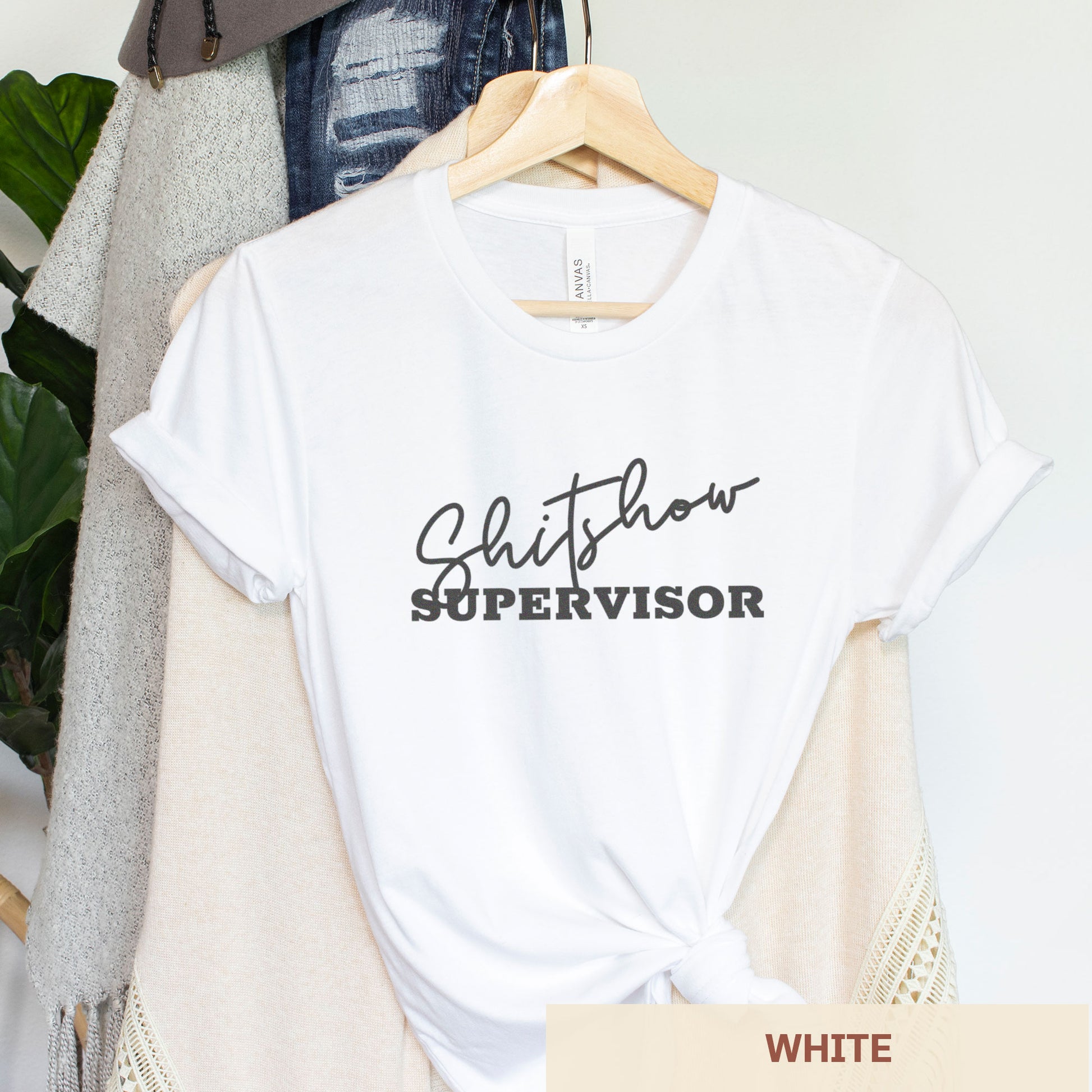 A hanging white Bella Canvas t-shirt featuring the text shitshow supervisor.