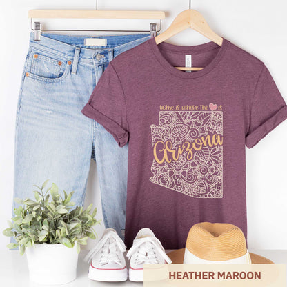 A hanging heather maroon Bella Canvas t-shirt featuring a mandala in the shape of Arizona with the words home is where the heart is.
