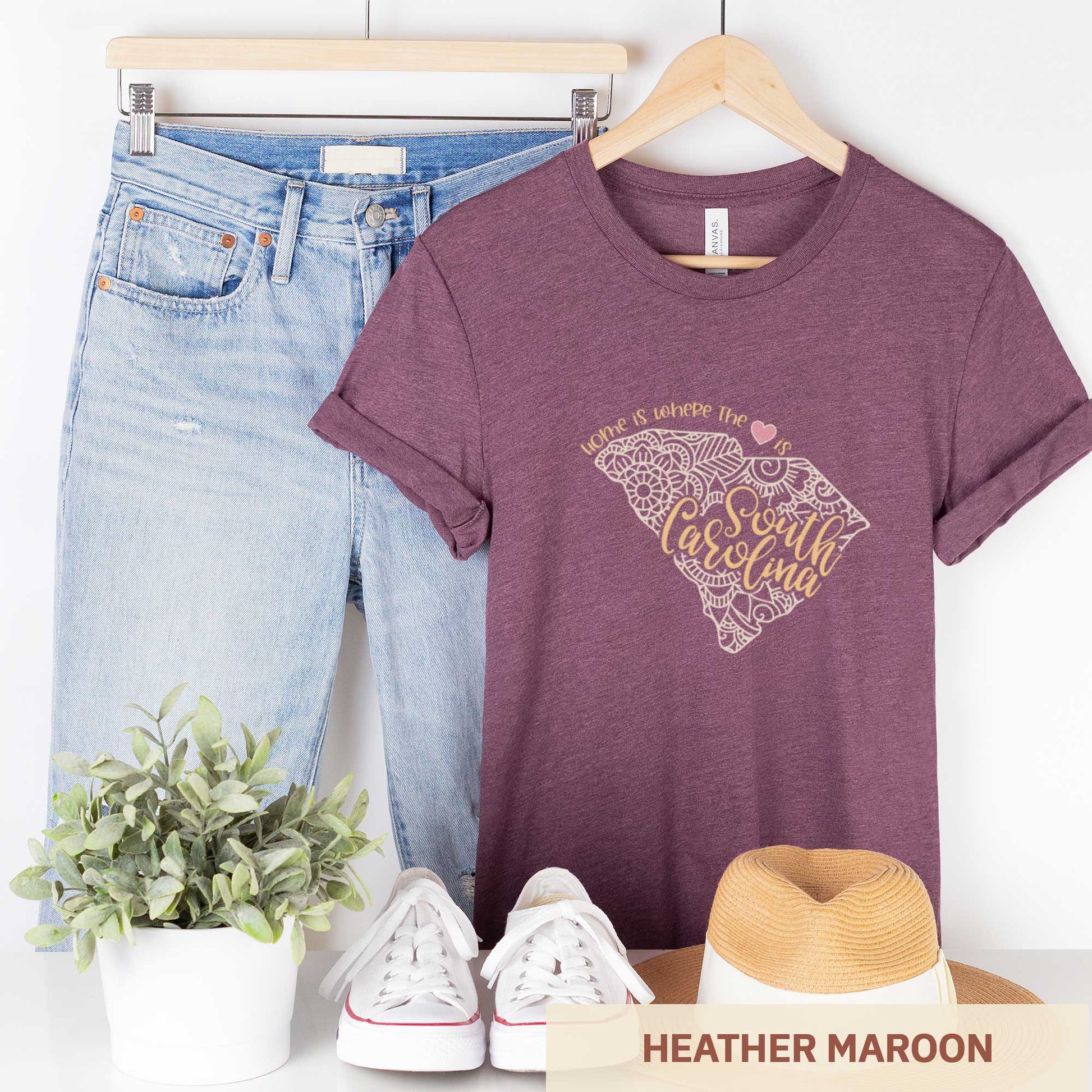 A hanging heather maroon Bella Canvas t-shirt featuring a mandala in the shape of South Carolina with the words home is where the heart is.