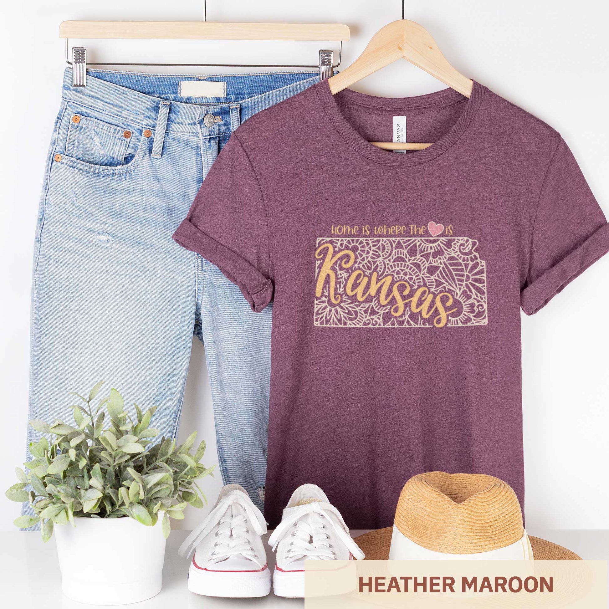A hanging heather maroon Bella Canvas t-shirt featuring a mandala in the shape of Kansas with the words home is where the heart is.