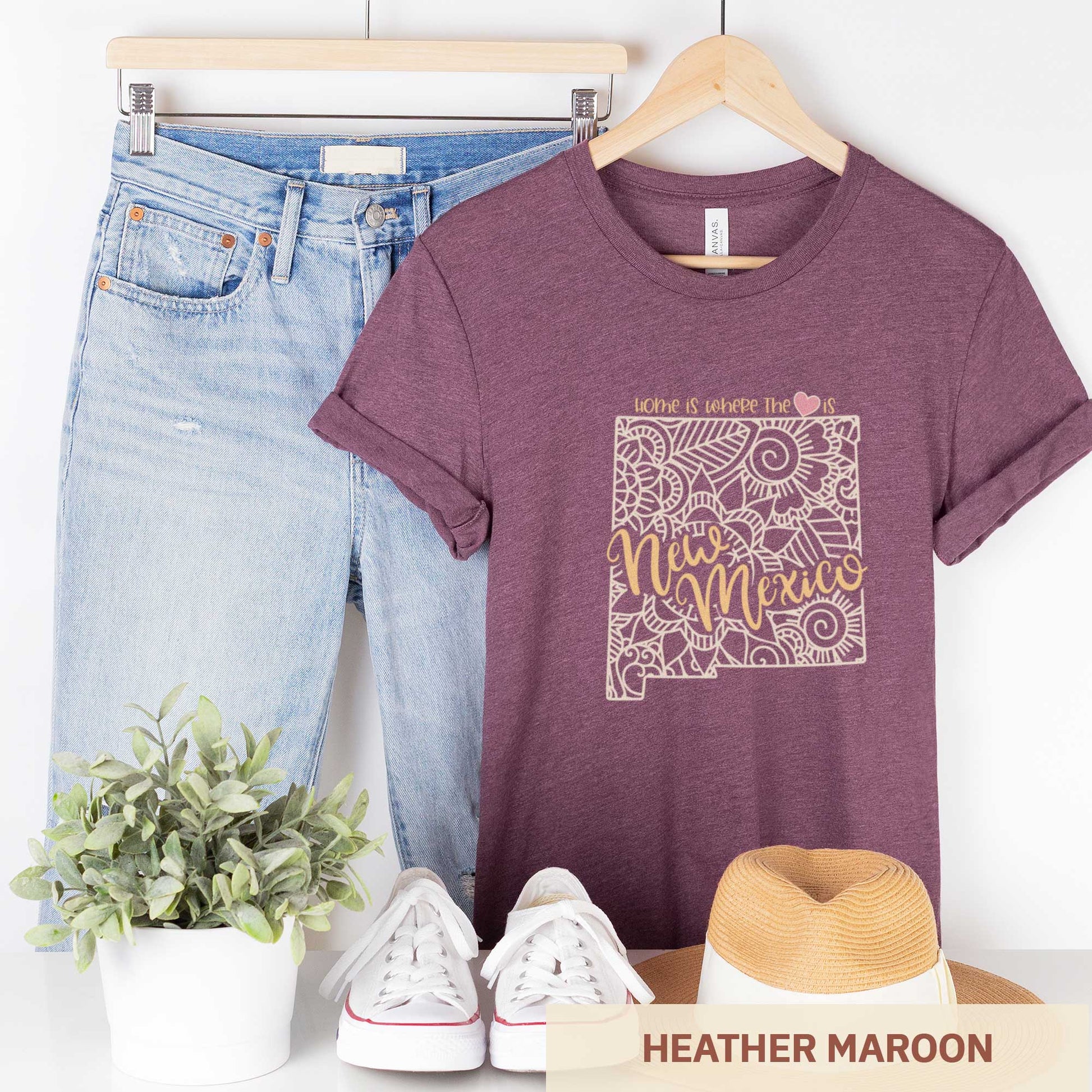 A hanging heather maroon Bella Canvas t-shirt featuring a mandala in the shape of New Mexico with the words home is where the heart is.