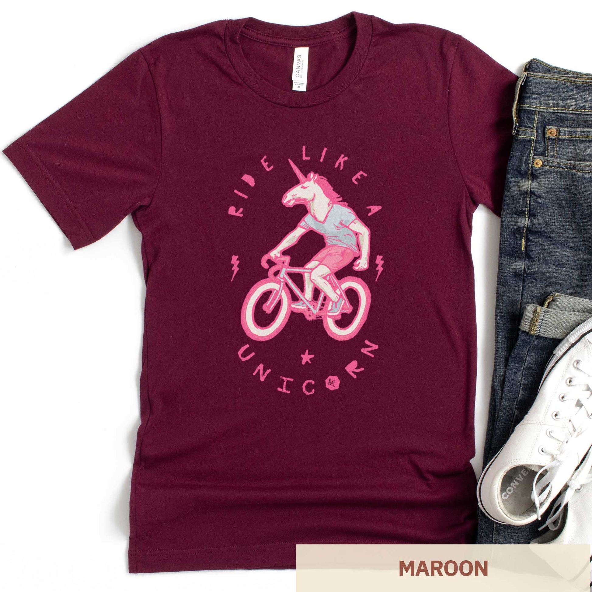 A maroon Bella Canvas t-shirt featuring a neon pink unicorn riding a bike and the words ride like a unicorn.