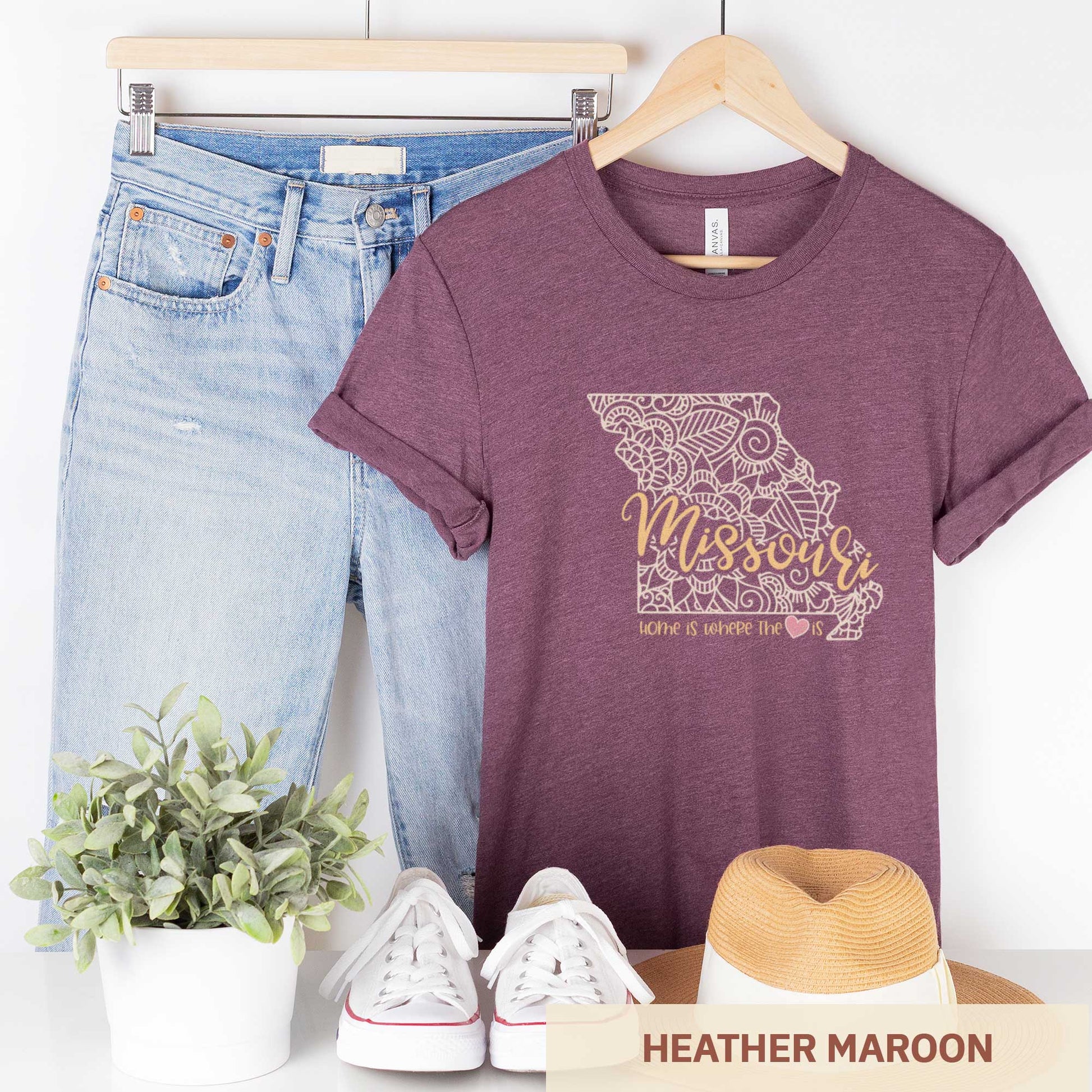 A hanging heather maroon Bella Canvas t-shirt featuring a mandala in the shape of Missouri with the words home is where the heart is.