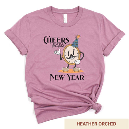 A heather orchid Bella Canvas t-shirt featuring a retro looking clock cartoon with a party hat next to the words Cheers to the New Year.