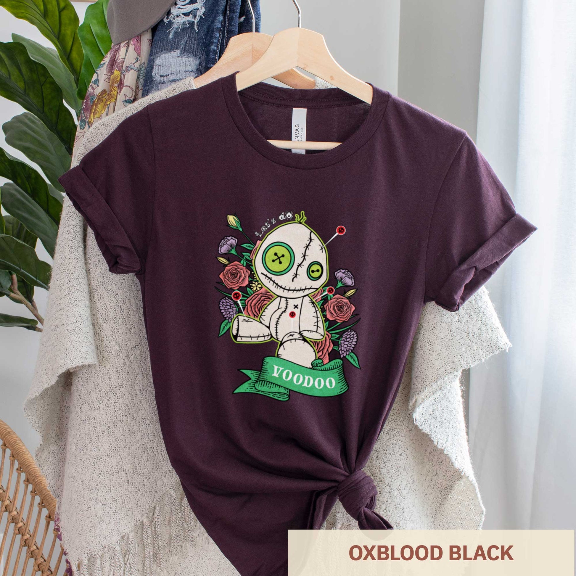 An oxblood black heather Bella Canvas t-shirt featuring a cartoon voodoo doll and flowers.