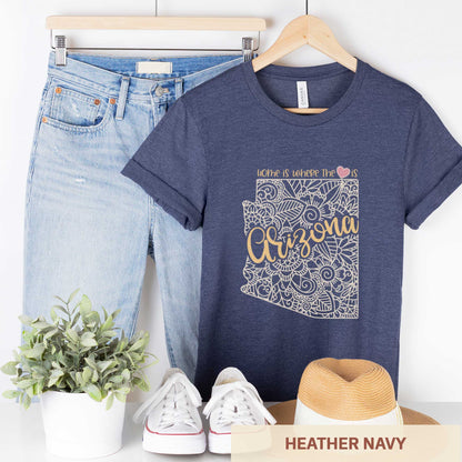 A hanging heather navy Bella Canvas t-shirt featuring a mandala in the shape of Arizona with the words home is where the heart is.