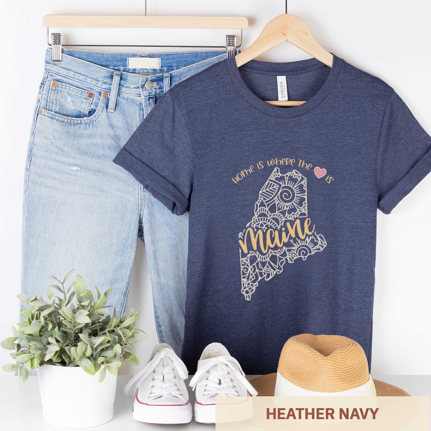 A hanging heather navy Bella Canvas t-shirt featuring a mandala in the shape of Maine with the words home is where the heart is.