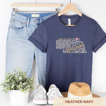 A hanging heather navy Bella Canvas t-shirt featuring a mandala in the shape of Nebraska with the words home is where the heart is.
