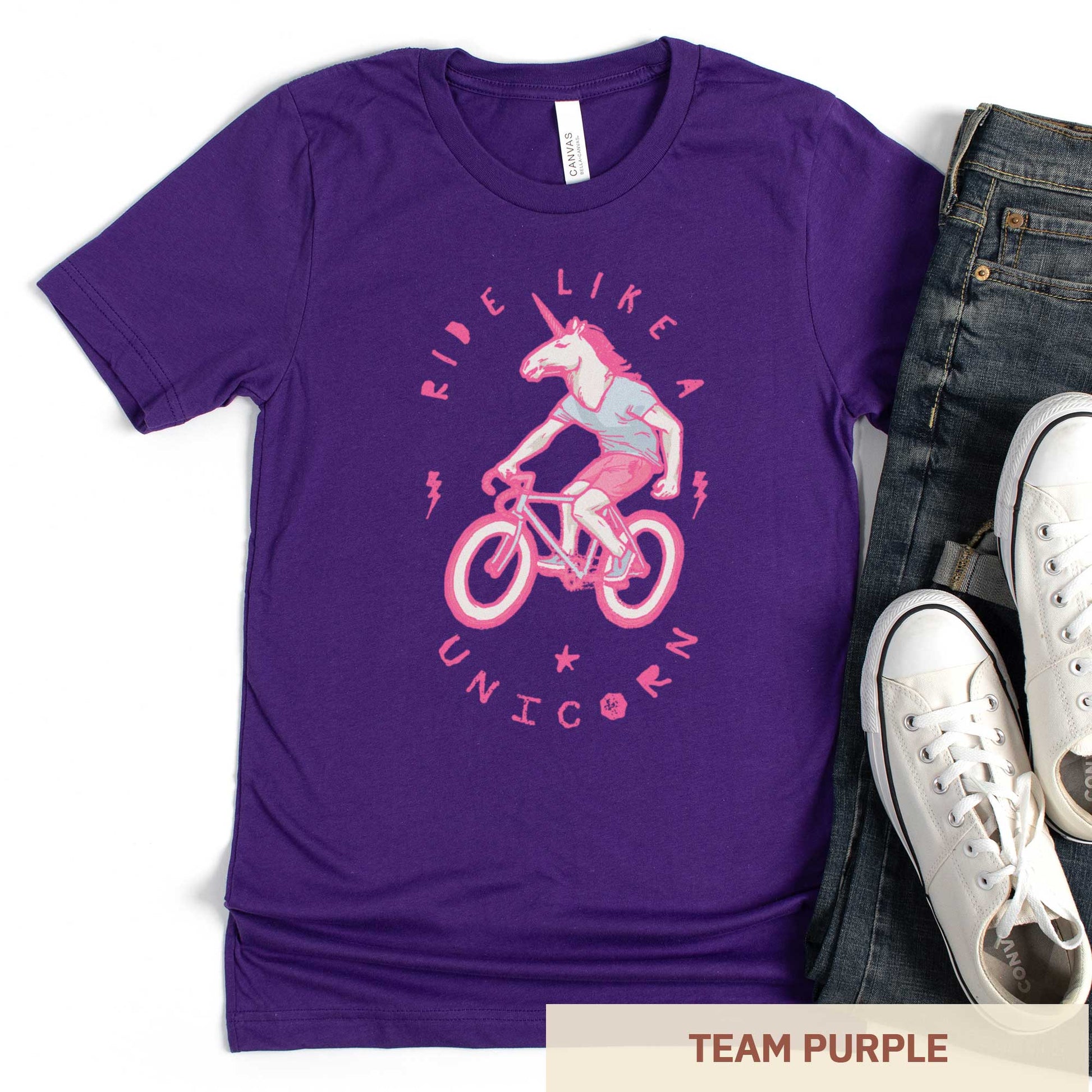 A team purple Bella Canvas t-shirt featuring a neon pink unicorn riding a bike and the words ride like a unicorn.