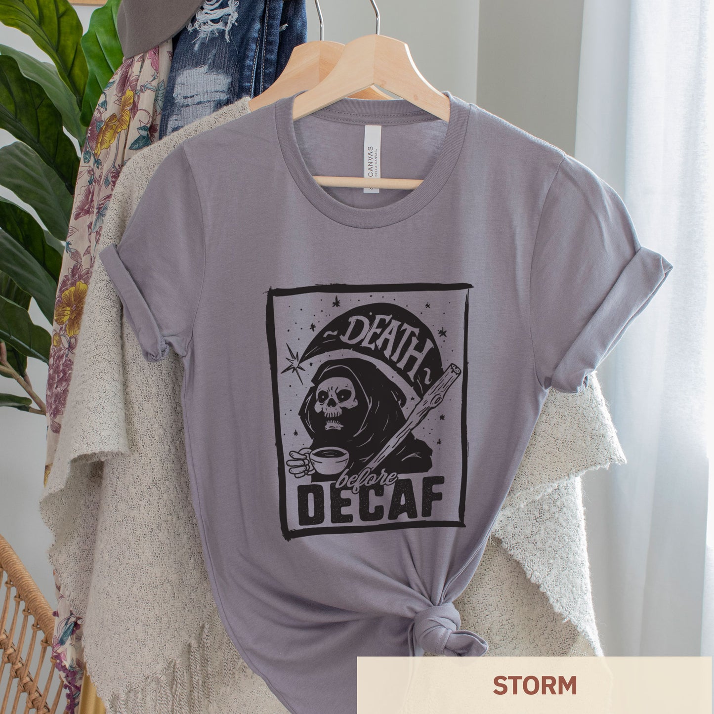 A hanging storm gray Bella Canvas t-shirt featuring the grim reaper drinking a cup of coffee with the words Death before decaf
