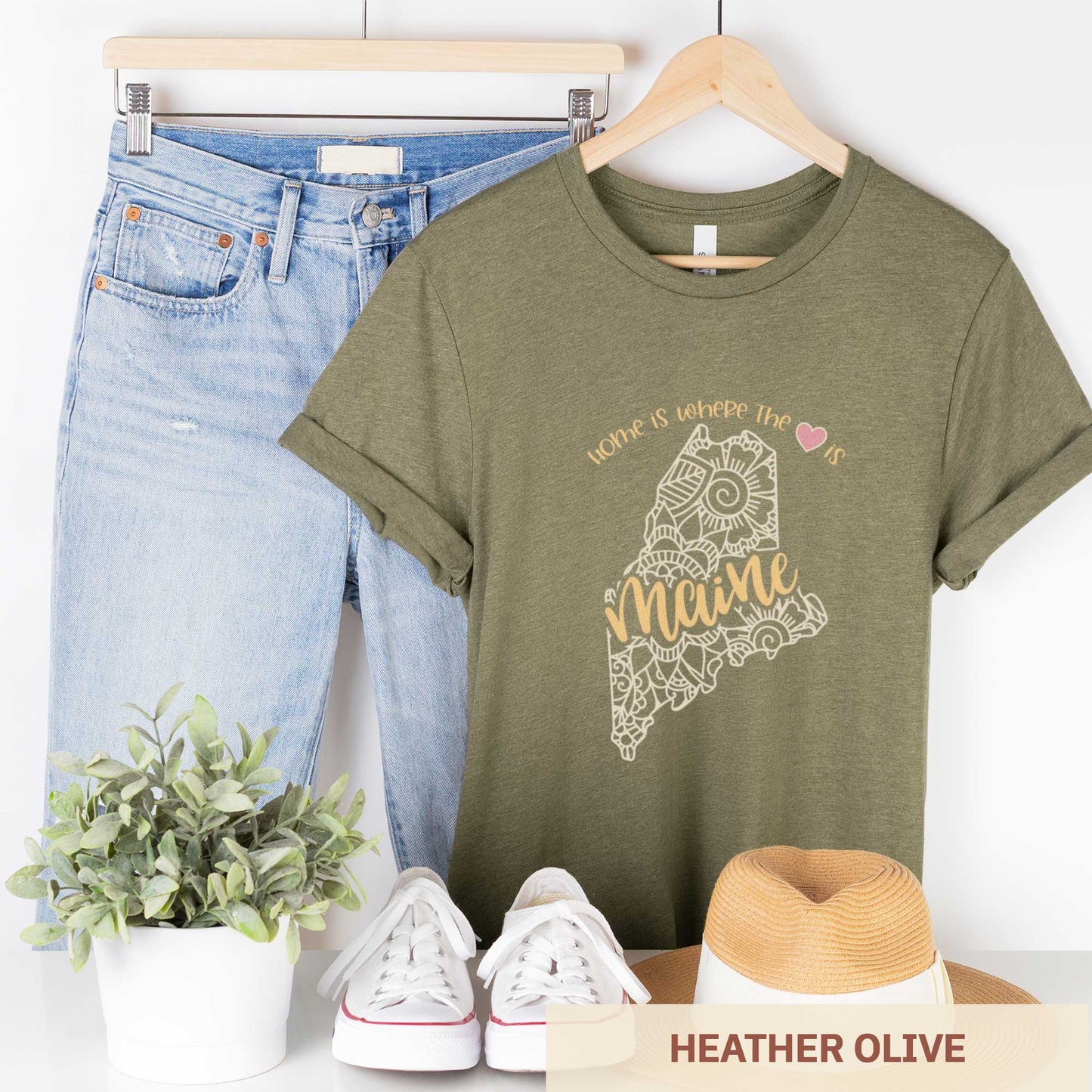 A hanging heather olive Bella Canvas t-shirt featuring a mandala in the shape of Maine with the words home is where the heart is.