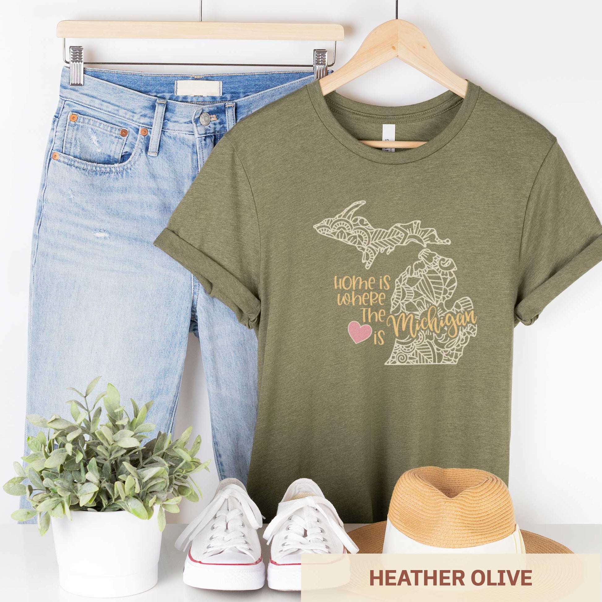 A hanging heather olive Bella Canvas t-shirt featuring a mandala in the shape of Michigan with the words home is where the heart is.