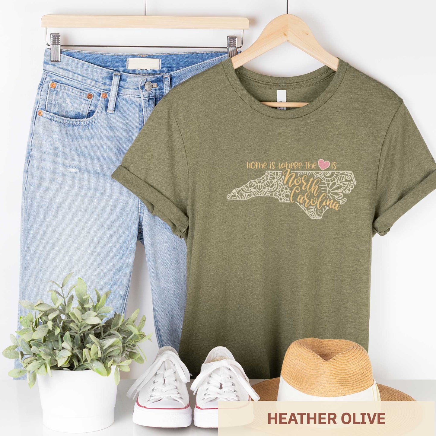 A hanging heather olive Bella Canvas t-shirt featuring a mandala in the shape of North Carolina with the words home is where the heart is.