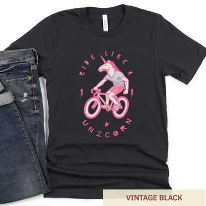 A vintage black Bella Canvas t-shirt featuring a neon pink unicorn riding a bike and the words ride like a unicorn.