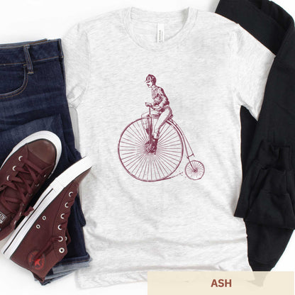 An ash Bella Canvas t-shirt featuring a Victorian man riding a penny farthing bicycle.